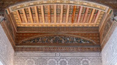 Painted Bahia Palace timber ceiling above a decorative frieze containing small human and animal figures.