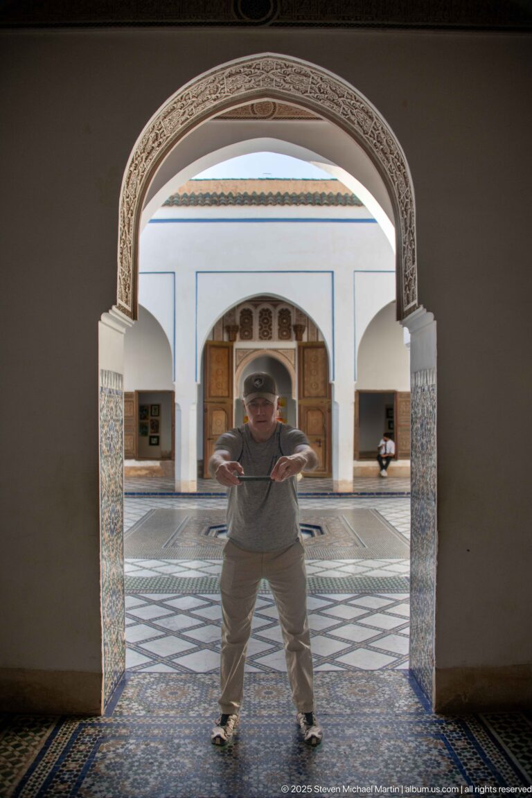 Visitor standing beneath a carved arch, with Bahia Palace courtyard paving and a small fountain visible behind.