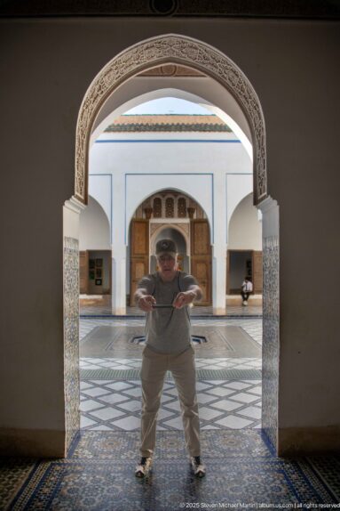 Visitor standing beneath a carved arch, with Bahia Palace courtyard paving and a small fountain visible behind.