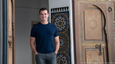 Visitor standing beside an open painted wooden Bahia Palace door, with colorful zellij mosaic visible behind.