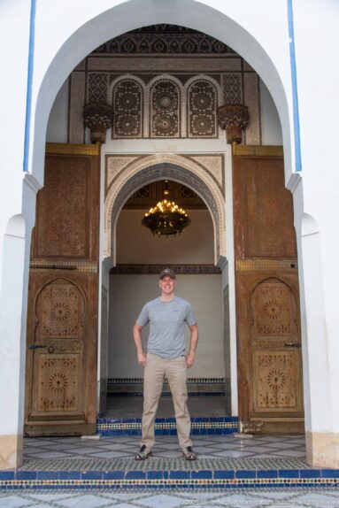 Visitor centered in a Bahia Palace doorway between carved wooden doors, beneath a hanging lamp and ornate arch.