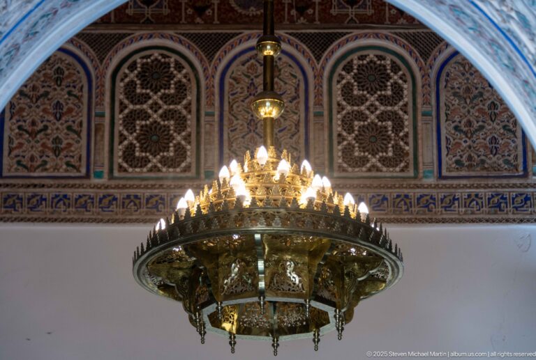 Large brass-colored Bahia Palace chandelier with many small bulbs hanging before painted and carved arched wall panels.
