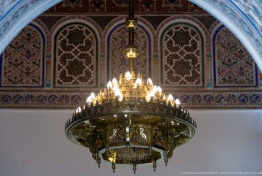 Large brass-colored Bahia Palace chandelier with many small bulbs hanging before painted and carved arched wall panels.