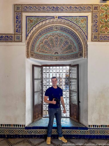 Visitor standing beneath a blue-green painted fan lunette and in front of a metal grille at Bahia Palace.