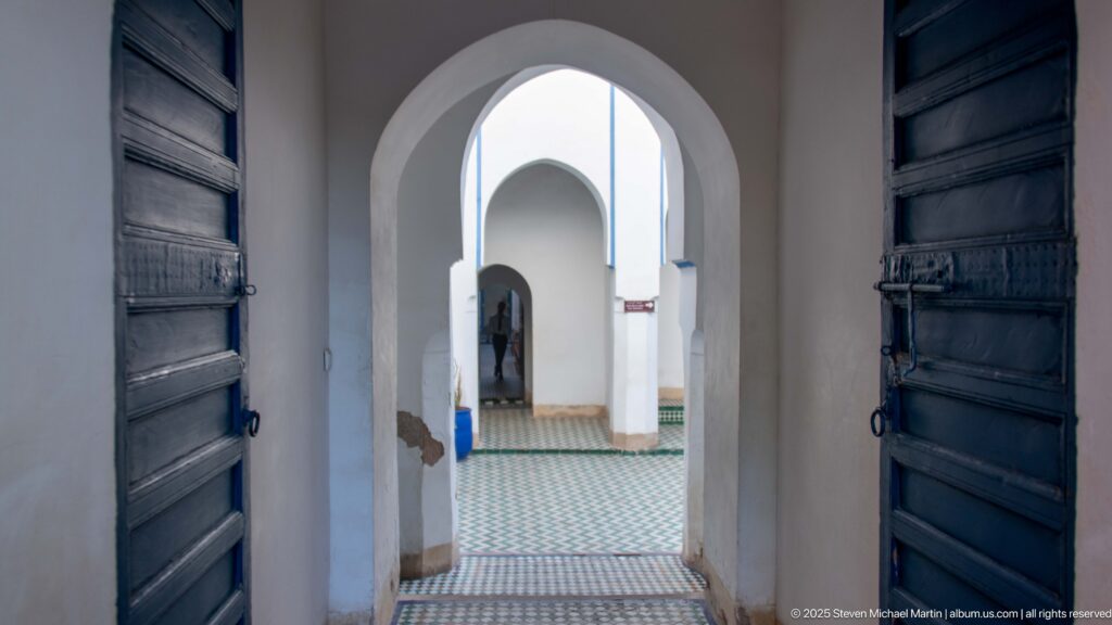 Open dark wooden doors framing a sequence of white arches and tiled flooring inside Bahia Palace.