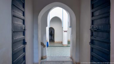 Open dark wooden doors framing a sequence of white arches and tiled flooring inside Bahia Palace.