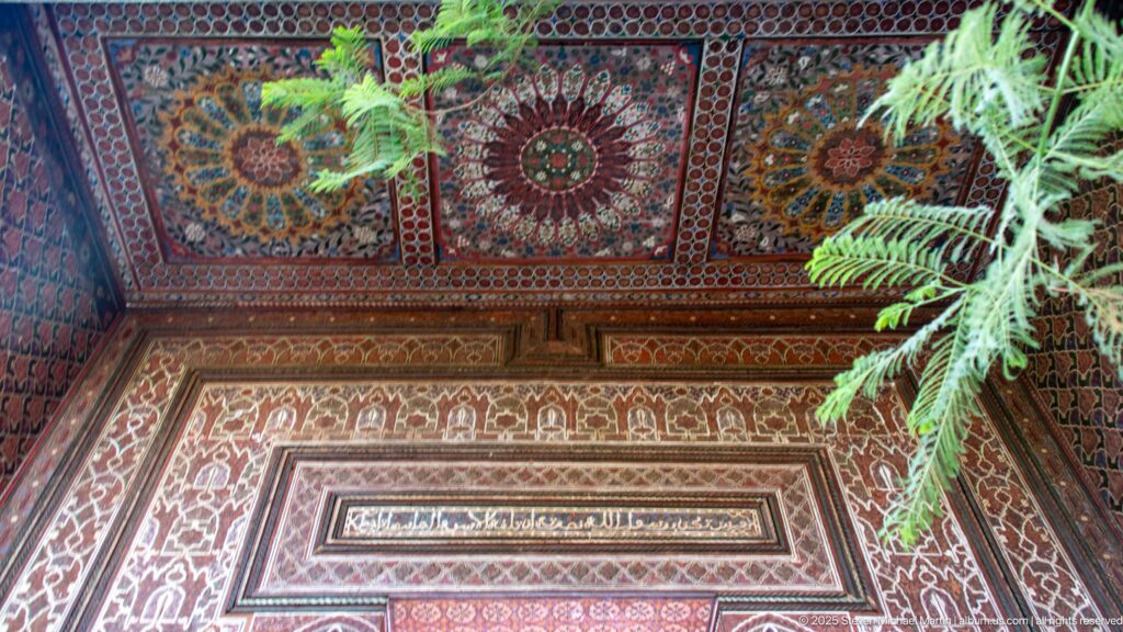 Polychrome Bahia Palace wooden ceiling corner with circular rosettes, floral patterns, framed central panel and green foliage.