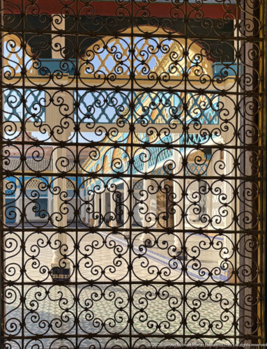 Black scrolling wrought-iron grille at Bahia Palace with blue-and-white courtyard architecture visible beyond.