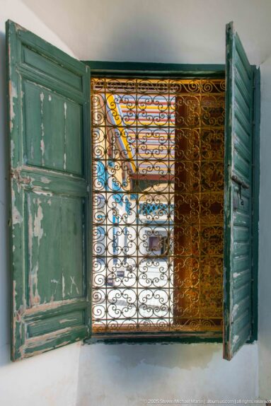 Weathered turquoise shutters opened around an ornate iron grille, with Bahia Palace courtyard visible through the screen.