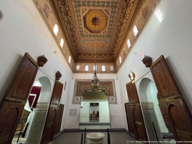 Long Bahia Palace Grand Riad hall with painted ceiling, open carved doors, hanging lantern and small central basin.