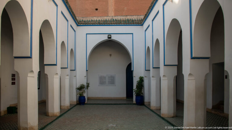 Small Bahia Palace courtyard with white pointed arches, blue trim, tiled paving and potted plants.