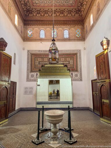 Centered Bahia Palace Grand Riad interior with stone basin, hanging lantern, open carved doors and painted decoration.