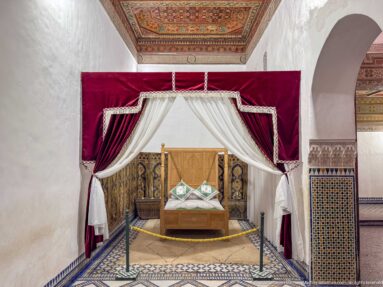 Wooden bed beneath red velvet and white curtains in a tiled, decorated room at Bahia Palace.