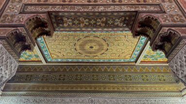 Polychrome Bahia Palace ceiling with radiating central medallion, carved brackets and pale plaster arches.