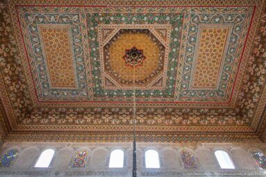 Painted Bahia Palace ceiling with central octagonal star medallion and colored-glass windows along the wall below.