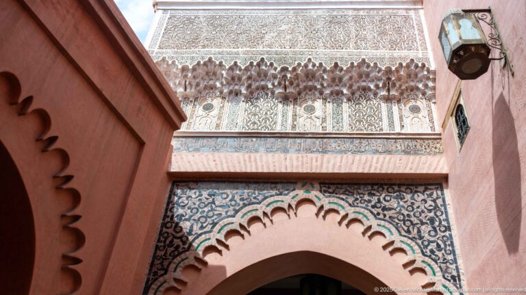 Upward view of an interior arch framed by blue-and-white zellij, carved stucco, and a projecting band of ornament.
