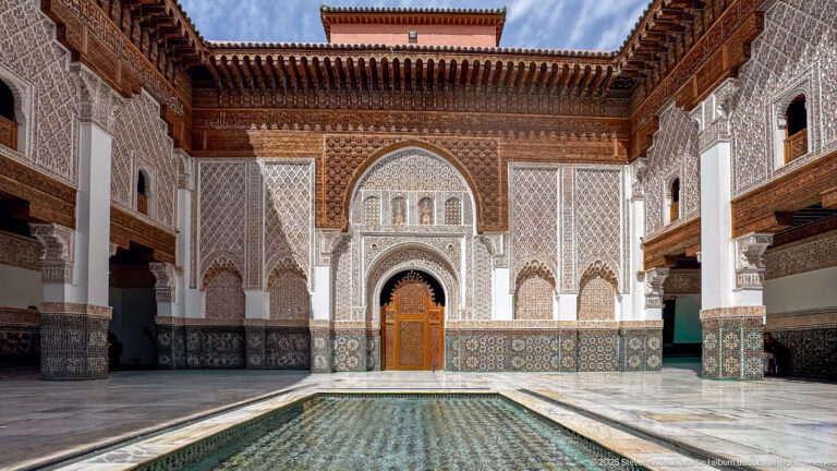 Rectangular courtyard pool centered on an ornate arched doorway, with white paving and decorated arcades on both sides.