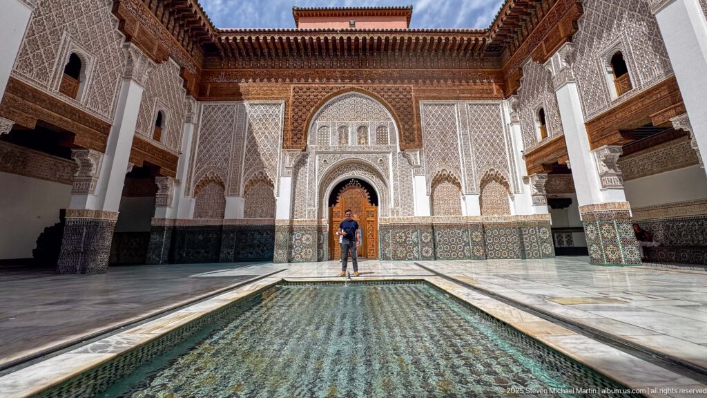 Low axial view along the rectangular courtyard pool toward a person standing before the ornate far doorway.