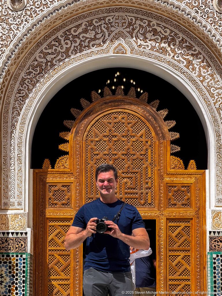Man holding a camera in front of carved wooden doors framed by pale stucco ornament and a narrow band of zellij.