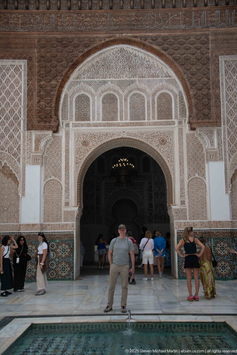 Large carved courtyard arch opening into a dark interior, with visitors gathered beneath it and the pool edge in the foreground.
