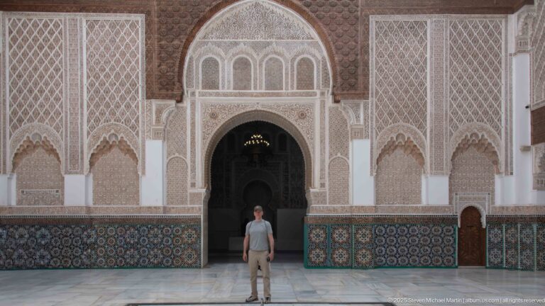 Single visitor centered beneath a tall decorated courtyard arch with carved stucco and zellij panels on both sides.