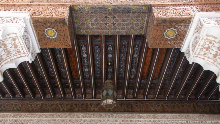 Upward view of a painted timber ceiling with dark beams, patterned panels, and a central hanging light between carved wall edges.