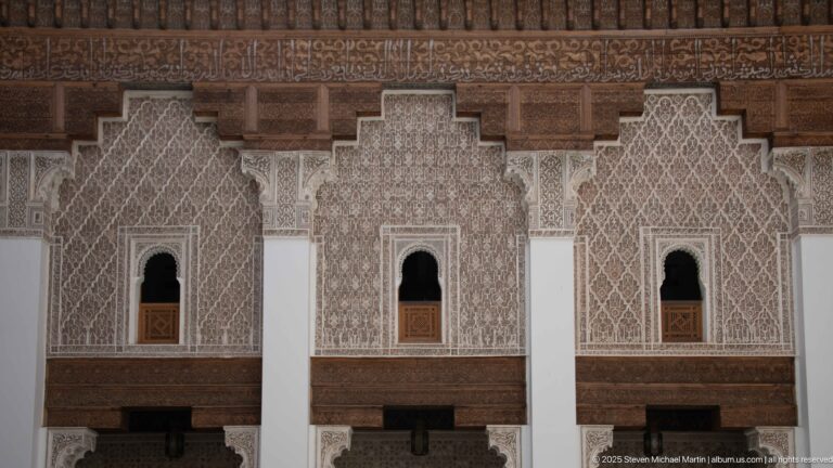 Three narrow upper-level openings set in dense carved stucco, separated by white piers beneath a band of dark wood.