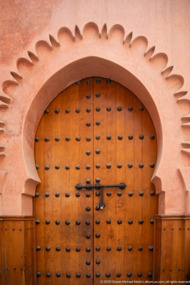 Large arched wooden door set in a scalloped plaster surround, covered with rows of dark metal studs and a central latch.
