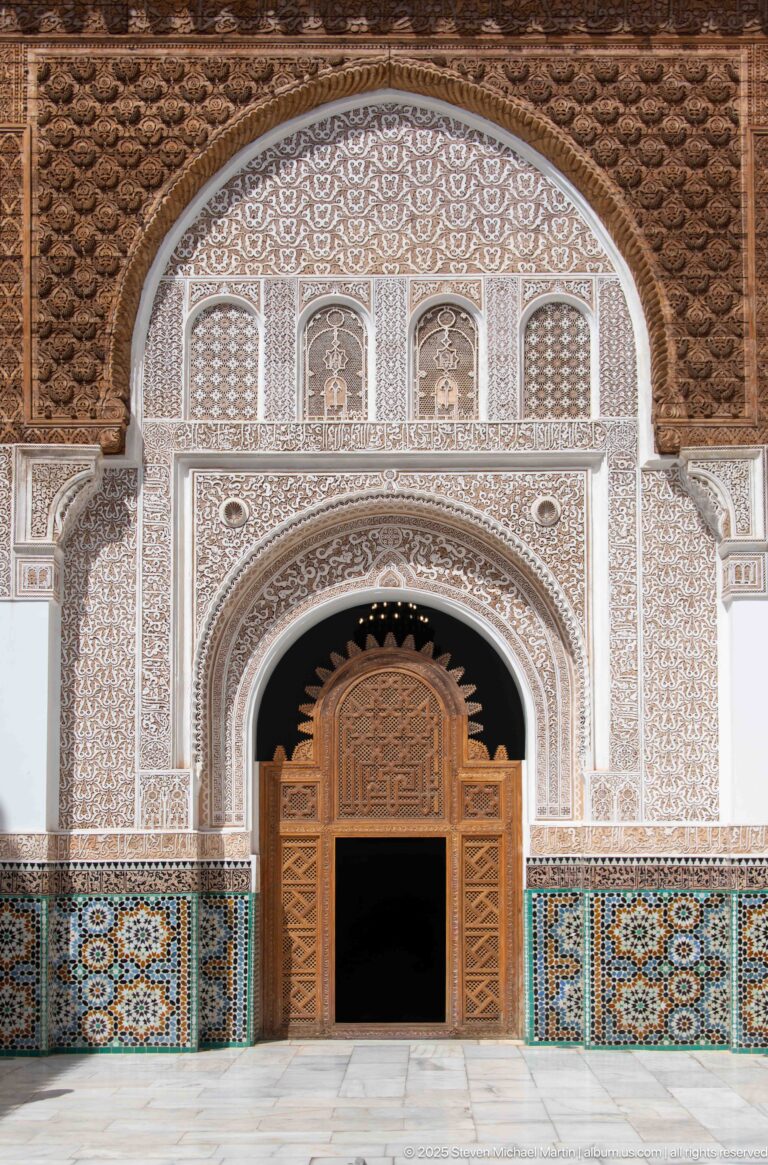 Open carved wooden doors in a richly decorated arch framed by pale stucco carving and multicolored zellij panels.