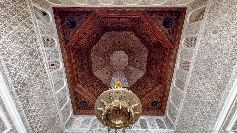 Upward view of a recessed polygonal wooden ceiling with central lantern, surrounded by pale carved walls and small screened openings.