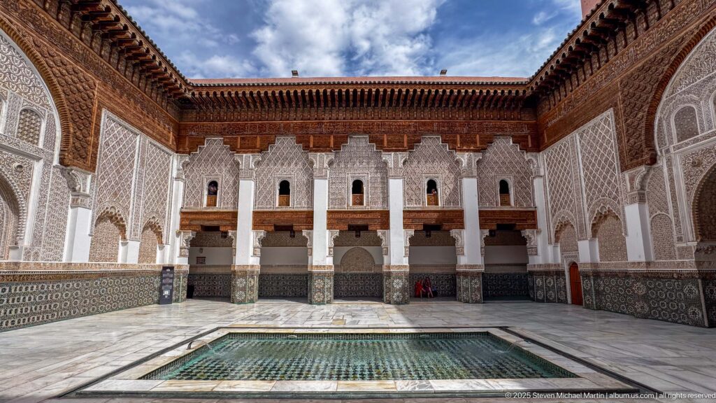 Wide courtyard side wall with repeated upper openings, shaded lower arcade, zellij panels, and the central pool in front.