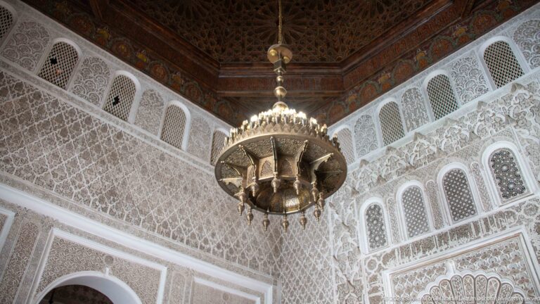 Large suspended metal lantern beneath a dark wooden ceiling, with carved pale walls, screened openings, and an arched niche.