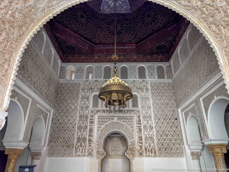 Frontal interior view with central arched niche, carved pale walls, dark wooden ceiling, hanging lantern, and side arches.