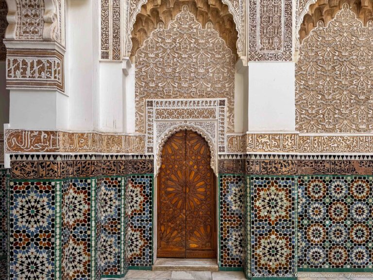 Small carved wooden pointed-arch door set within multicolored zellij, with pale carved stucco panels rising above it.