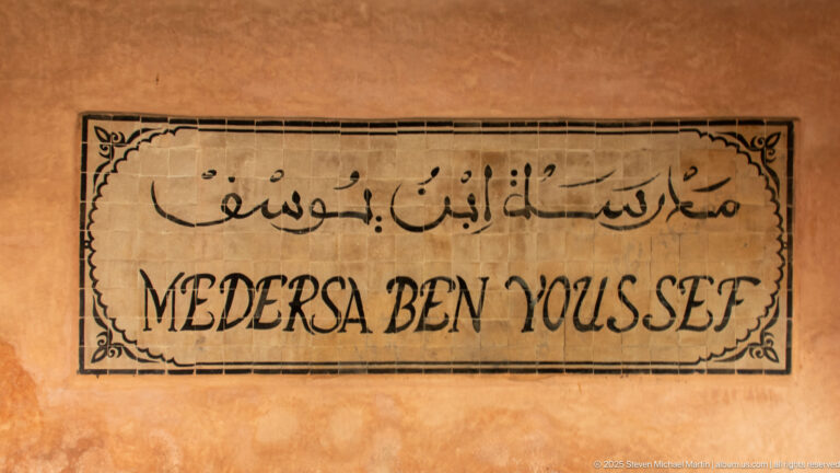 Rectangular wall sign on ochre plaster reading “MEDERSA BEN YOUSSEF” beneath Arabic script, with a thin decorative border.