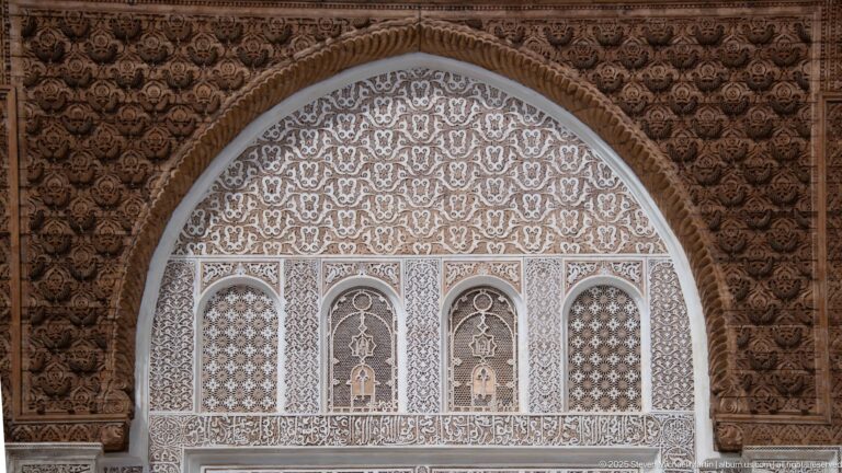 Large upper arched wall panel with four small screened openings, pale carved ornament, and a dark carved wooden band above.