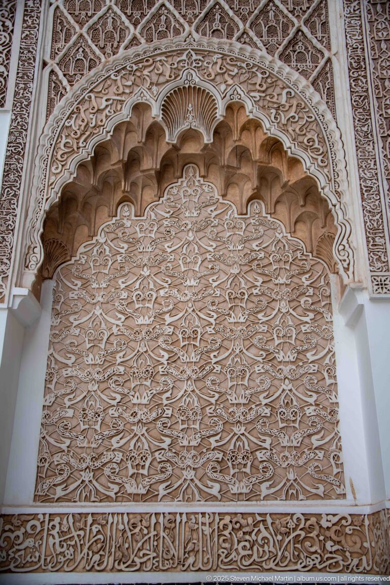 Cream carved stucco arch with scalloped inner molding, dense arabesques, and Arabic calligraphy.
