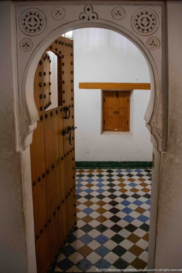 Open studded wooden door in a scalloped arch leading to a white room with geometric tile floor.