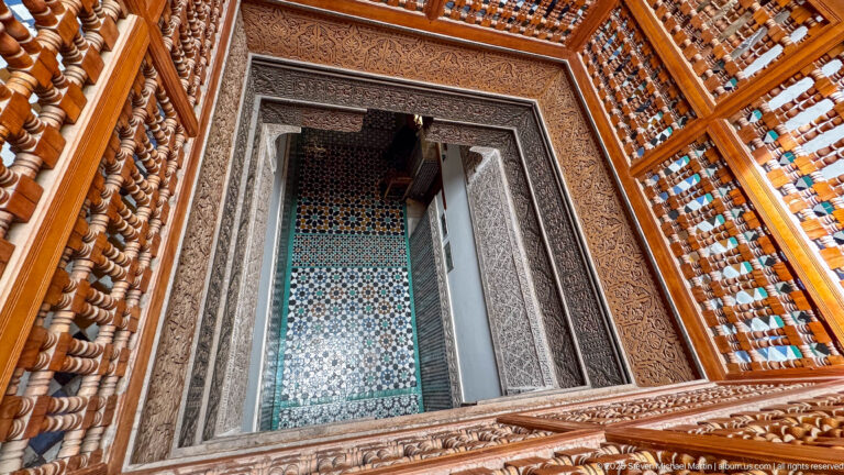 Oblique view through carved wooden balustrades toward a stucco-framed opening and geometric tilework.