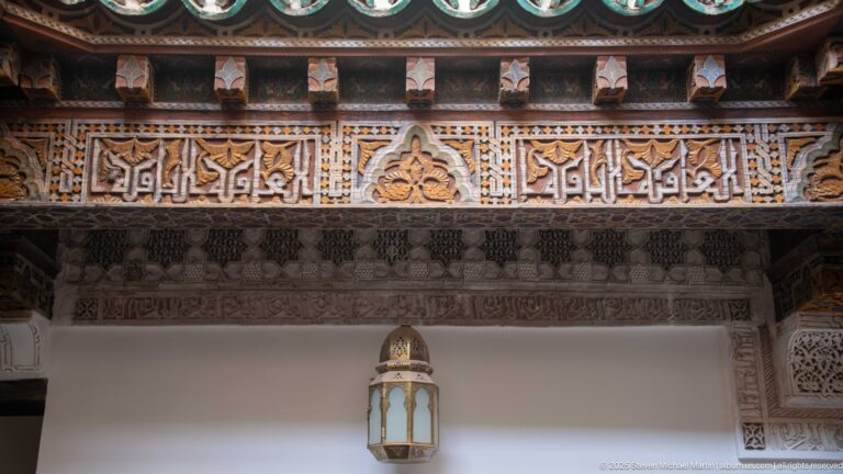 Arabic calligraphic frieze and painted timber ornament above a white wall with a hanging lantern.