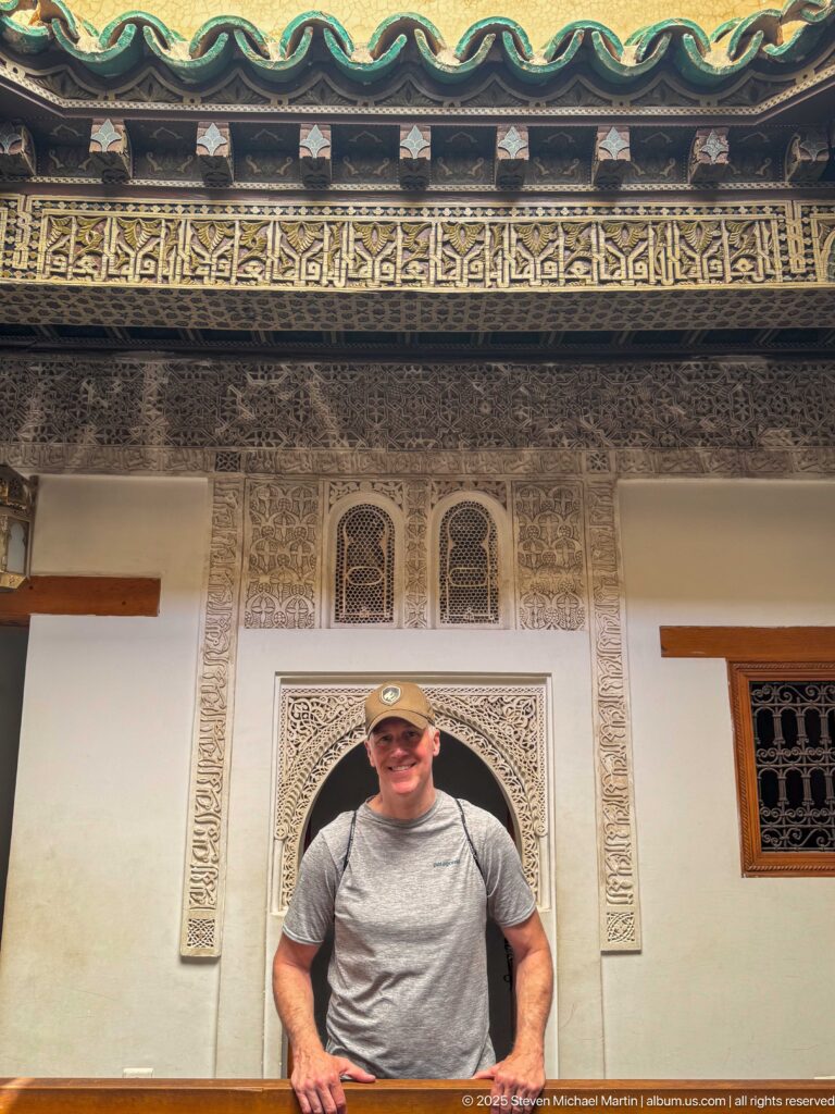 Smiling visitor standing before carved plaster panels, a small arch, and an Arabic calligraphic frieze.