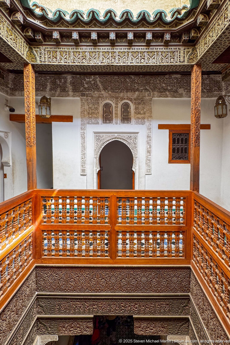 Wide upper-gallery view with wooden balustrades, carved plaster walls, central arch, and painted eaves.