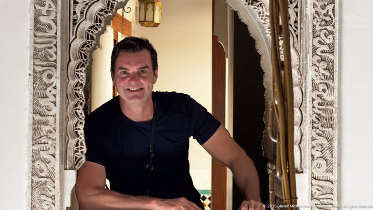 Visitor in dark shirt framed by an ornate carved stucco opening, with a lantern and doorway behind.