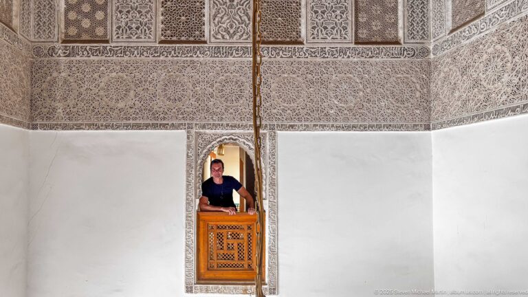 Wide carved plaster wall with a small central arched opening where a visitor stands behind a wooden lattice.