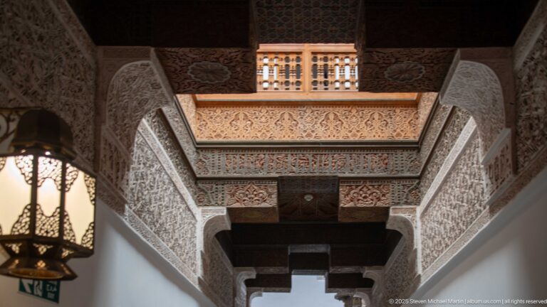 Low-angle view along a passage ceiling with dark wood beams, carved decorative bands, and a hanging metal lantern at left.