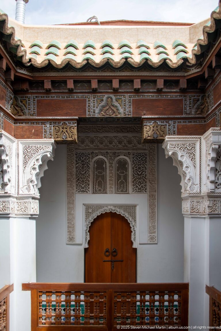 Central wooden double door beneath carved plaster panels and painted timber eaves, with gallery railing in front.
