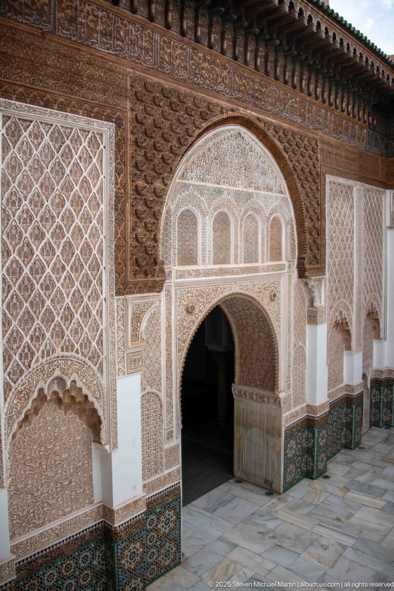 Oblique courtyard wall with large horseshoe doorway, carved stucco panels, and green-blue zellij along the base.