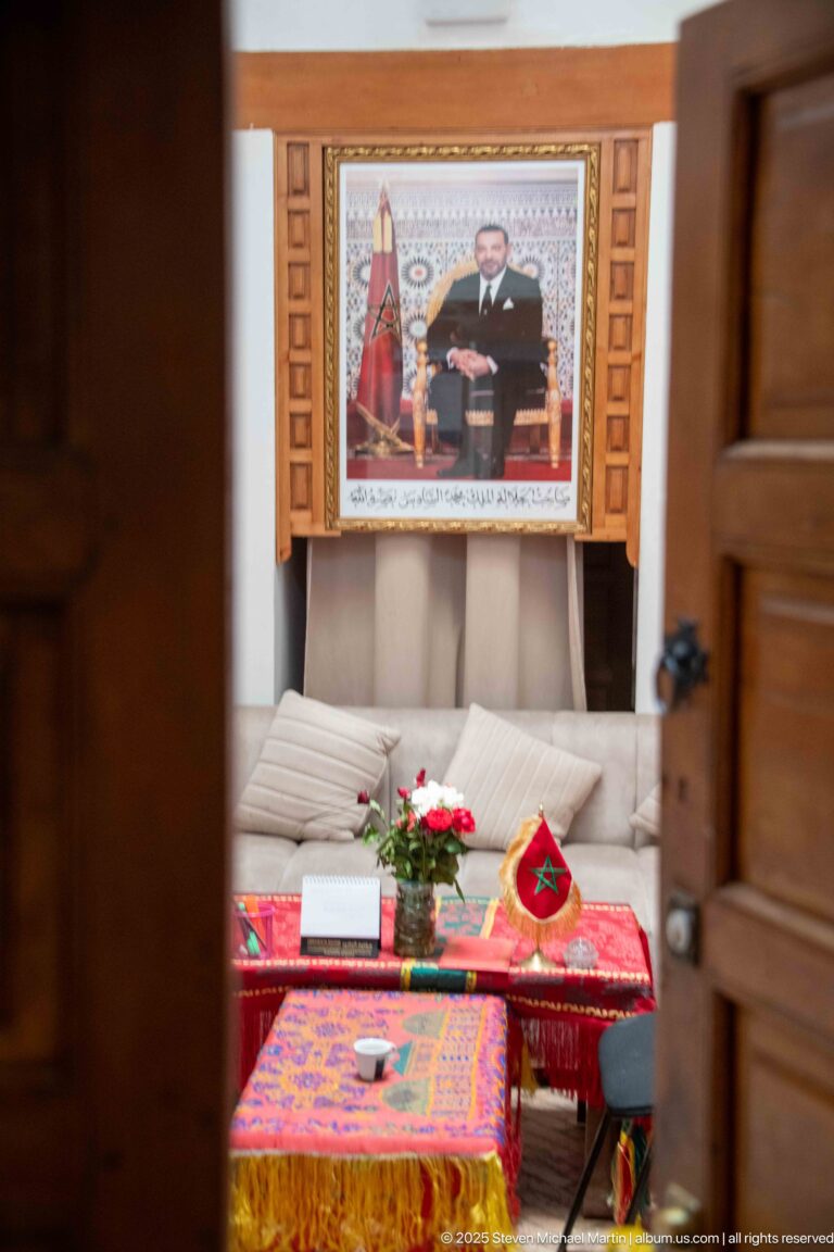 Partly open wooden door revealing a furnished room with patterned textiles, flowers, Moroccan symbols, and a framed portrait.