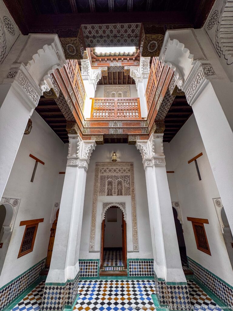 Interior court with tall white piers, geometric tile floor, zellij dado, carved doorway, and wooden gallery above.