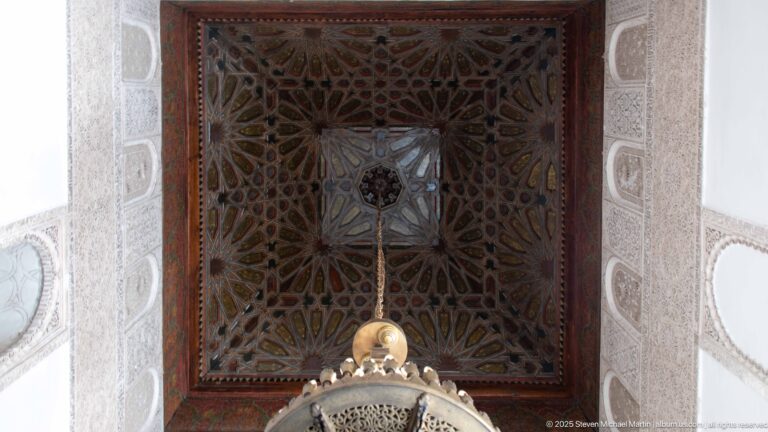Square decorative wooden ceiling with interlaced geometric star patterns, framed by pale carved walls and a hanging lantern below.